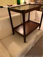 Photo of wood tea trolley showing front side view with caster wheels and two shelves on a white couch.