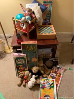 Wooden doll crib with various plush teddy bears and dolls, vintage games arranged in front including Plunk, Crazy Maze, Mercury Maze, Kaplow, and paperback books visible. Baby bath tub and baby bath seat visible. Lot shown on wooden table.