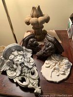 Five decorative casting pieces shown on a table including two cherub wall sconces, an ornate face casting, a flower casting, and a finial-style piece.