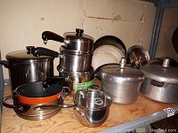 Overall view of various cookware including pots, pans, lids and stacked pressure cookers on wooden surface.