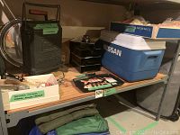 Shelf with black camp stove, blue Nissan cooler, small utensil boxes and green and blue folded camping mats below