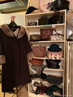 Full view of vintage wool coat with fur collar and cuffs. Various vintage handbags, hats, and belts arranged on shelves behind.