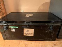 Photo of entire closed vintage black metal travel trunk showing rectangular shape and sticker with dimension notes on front.