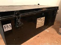 Black rectangular metal travel trunk with visible wear and scratches on surface, metal latches on front panel, handwritten paper label pasted on trunk front.