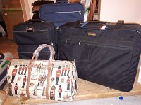 Photo showing four black luggage pieces including a large duffel bag and three suitcases, alongside a decorative handbag with British royal guards print.