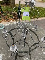 View of metal chandelier with 5 curved arms, each with a bulb holder and glass bobeche, on a glass table outdoors. Silver finish and hanging glass accents visible.