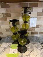 Three green graduated glass candle holders with dark metal tops and bases, two with multi-color butterfly charms hanging from green bead strings, arranged on a marble surface against a tile backsplash.