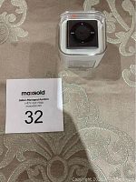Photo of unopened Apple iPod Shuffle 2GB black in original clear square plastic packaging on a patterned surface alongside a MaxSold auction card with number 32.