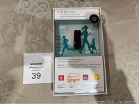 Front view of new Fitbit One activity tracker in retail packaging showing product and illustrations of usage.