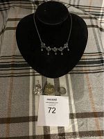 Black necklace display bust with silver-tone floral necklace and four assorted pendants and brooch placed below it on a plaid fabric background.