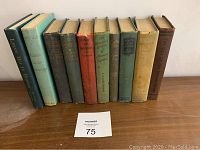 Photo showing a row of 11 antique hardcover books with varied colors and visible titles on spines, arranged on a wood surface with a lot number card '75' in front.