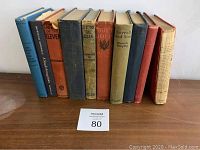 Nine antique books standing upright, showing spines with titles and signs of wear.