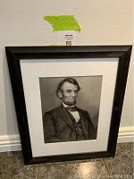 Frontal view of the framed Abraham Lincoln picture in black and white with white matting and black frame on floor against a wall.