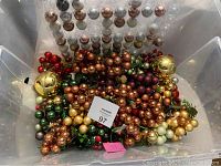 Clear plastic container filled with assorted shiny and matte Christmas baubles including gold, bronze, green, red, and pearl white, along with artificial berry clusters.