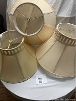 Group photo of 3 beige/tan fabric lamp shades with wired frames, showing different shapes and sizes, placed on a white surface.
