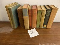 Photo showing ten antique hardcover books arranged upright on a wooden surface with a lot number card (108) in front.