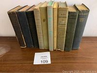 Lot of nine antique hardcover books standing upright with visible wear and tear, resting on a wooden surface with MaxSold lot number 109.