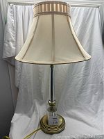 Full view of the lamp showing the beige fabric shade, brass and glass body, and round brass base.
