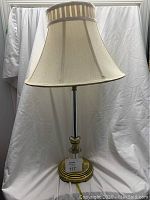 Full view of lamp with shade, fabric lampshade beige with striped trim, brass and glass base, placed on white draped background.