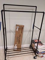 Full view of the assembled black metal rolling clothes rack with boxed rack behind it.