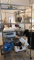 Metal shelving unit with KitchenAid stand mixer on top shelf, electric Nesco 18 roaster on middle shelf, Faberware grill and Black & Decker boxed food processor on bottom shelf. Shows overall lot contents.
