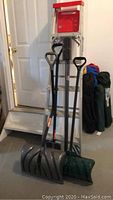 Photo of aluminum step ladder with red top and three shovels leaning against a wall with camping chairs wrapped in bags to the side.