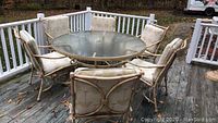 View of round patio table with six cushioned metal chairs arranged on wooden deck, daylight outdoor setting showing some dirt and leaves on deck.