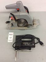 Photo showing side view of circular saw with gray plastic housing and Black & Decker jig saw with black housing in front, both with power cords.