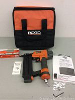 The Ridgid 18 gauge brad nailer lying flat on a surface beside a soft carrying case, two boxes of nails, a user manual, and a small plastic bag containing accessories.