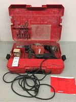 Open red plastic carrying case showing Hilti TE15 drill, side handle, cord, bits holder and instruction manual.