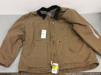 Front view of brown Carhartt work coat with black collar, zipped front, and pockets, showing tags attached.