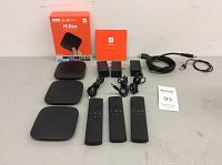 Set of three Xiaomi Mi Box 4K streaming devices shown with three remote controls, three power supplies, one HDMI cable, one micro USB cable, original box for one device, and some coupons.