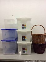Overall lot showing three white TUFF storage bins stacked, two clear plastic bins with blue lids stacked on the left, and two woven baskets on the right (brown with handle and dark purple).