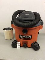 Front view of Ridgid orange shop vac showing black hose wrapped on top and one new cylindrical pleated filter standing beside it. Black plastic casters visible on base.