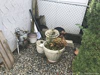Assorted plastic plant pots grouped on gravel outside near a water meter and fence. Large cream-colored pot contains a live plant with reddish leaves. Other pots in grey, purple, and black with varied sizes are around it. Photo taken for detailed inventory of plant pots and plant condition.