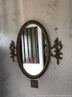 Oval plastic framed decorative wall mirror with two ornate decorative sconces mounted on wall beside it.