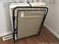 Metal folding cot frame folded vertically, black frame with elastic cords, stored against wall.