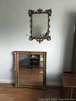 Two ornate framed wall mirrors, one larger plastic framed in dark gold and one larger wooden framed with baroque style, placed against light gray wall.