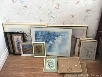 Wide shot of 14 different framed prints arranged on floor, mostly floral and some scenic, various frame colors and sizes from 5x8 to 19x16 inches