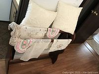 Wooden quilt rack with crocheted cream throw pillows and assorted crocheted and embroidered doilies, some with pink and green accents, hanging on it.