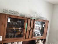 Wide view of glassware on top of and inside wooden cabinet showing multiple glass items including bowls, glasses, pitchers, and plates.