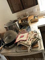 Large stainless steel stock pot, aprons, oven mitts, and some pans arranged on kitchen counter.