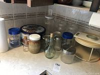 Photo shows assorted glass jars, canisters, a plastic juice jug, a garlic saver, and a two-tiered cream-colored plastic cake stand with clear dome lid on a kitchen counter.