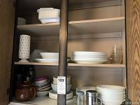 Wooden cabinet with white Corelle plates and bowls, glass bowls, metal coffee pot and pots on shelves.
