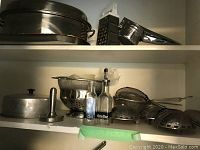 Multiple stainless steel kitchen items including roaster with lid, colander, sieves, glass oil and vinegar bottles, and kitchen tools on shelves.