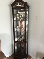 Full view of the wooden curio cabinet with three glass shelves containing decorative items, glass on front and sides, dark wood frame.