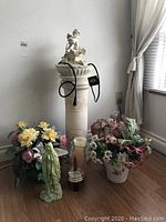 Photo showing the indoor fountain with cherub statue on top, pedestal base, power cord, and surrounding artificial floral bouquets.