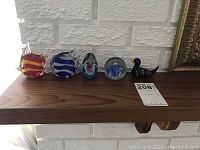 Five glass paperweights on a wooden shelf next to a white brick wall and auction lot tag visible