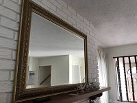Mirror hanging on white brick fireplace wall above wooden shelf. The mirror has a decorative gold-tone carved frame.