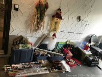 Wide view of assorted Christmas decorations on floor and stacked in boxes and bins, showing various items including a Santa face figure, greenery, and rolls of wrapping paper or light wrap.